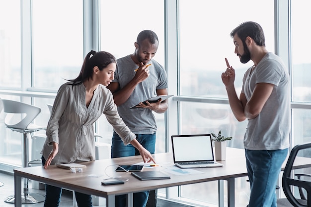 Groep aantrekkelijke multiraciale zakenmensen die samenwerken in een creatief kantoor terwijl ze in de buurt van het houten bureau staan, vriendelijk multi-etnisch zakenteam dat zakelijke problemen bespreekt tijdens de vergadering