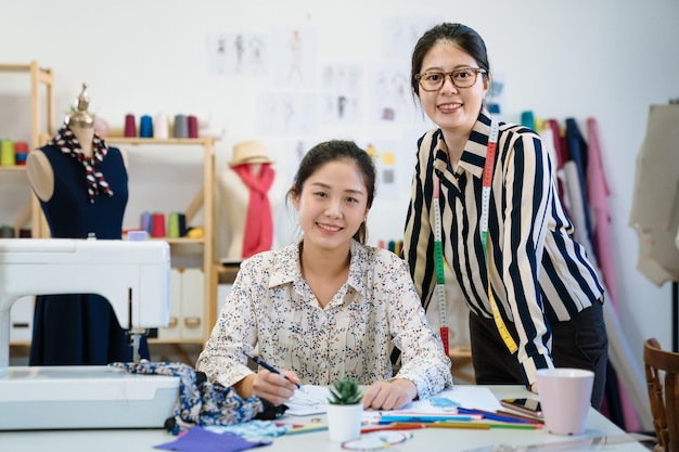 groep aziatische japanse glimlachende modeontwerpers vrouwen die werken aan een bureau en gezichtscamera. twee naaisters in creatief team. zelfverzekerde jonge dames collega's in gedeeld kantoor samen brainstormen.