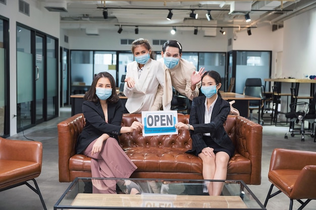 Groep aziatische zakenmensen die een gezichtsmasker dragen, zitten en houden heropenen en houden papier voor sociale afstand in een modern kantoor
