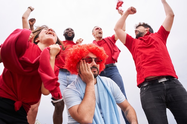 Groep fans van een voetbalteam op de tribunes die rode shirts dragen met een fan van het andere team