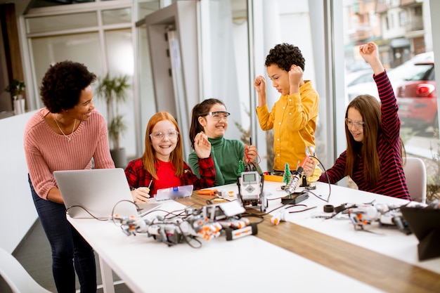 Groep gelukkige jonge geitjes met hun Afrikaanse Amerikaanse vrouwelijke wetenschapsleraar met laptop die elektrisch speelgoed en robots programmeren bij robotica-klas