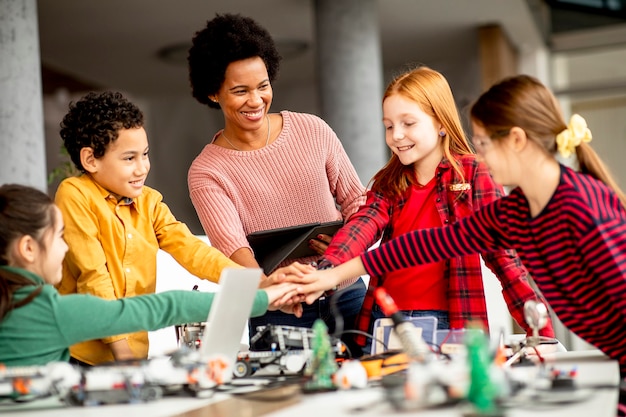 Groep gelukkige jonge geitjes met hun Afrikaanse Amerikaanse vrouwelijke wetenschapsleraar met laptop die elektrisch speelgoed en robots programmeren bij robotica-klas