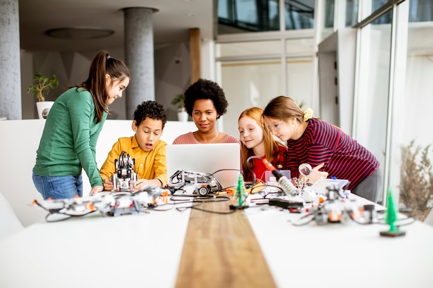 Groep gelukkige jonge geitjes met hun Afrikaanse Amerikaanse vrouwelijke wetenschapsleraar met laptop die elektrisch speelgoed en robots programmeren bij robotica-klas