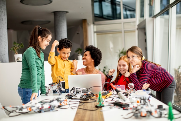 Groep gelukkige jonge geitjes met hun Afrikaanse Amerikaanse vrouwelijke wetenschapsleraar met laptop die elektrisch speelgoed en robots programmeren bij robotica-klas