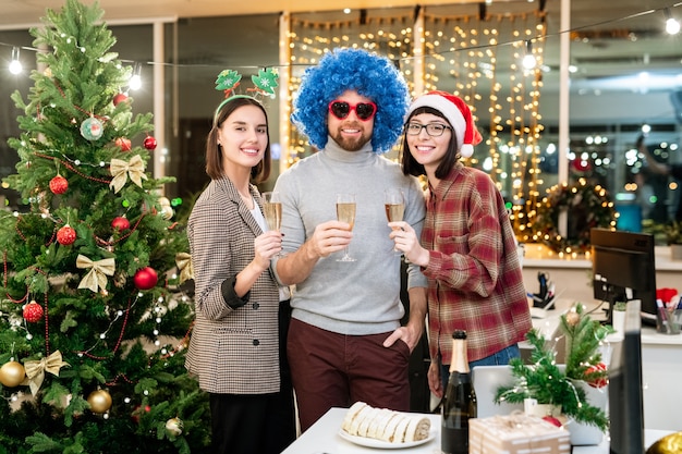 Groep gelukkige jonge ondernemers roosteren voor Kerstmis met fluiten champagne terwijl u geniet van feest in kantoor