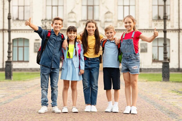 Foto groep gelukkige multi-etnische kinderen met rugzakken die buiten poseren en duimen omhoog laten zien