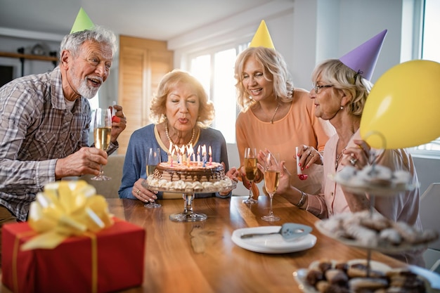 Groep gelukkige senioren die plezier hebben op verjaardagsfeestje terwijl vrouw thuis kaarsen blaast op verjaardagstaart