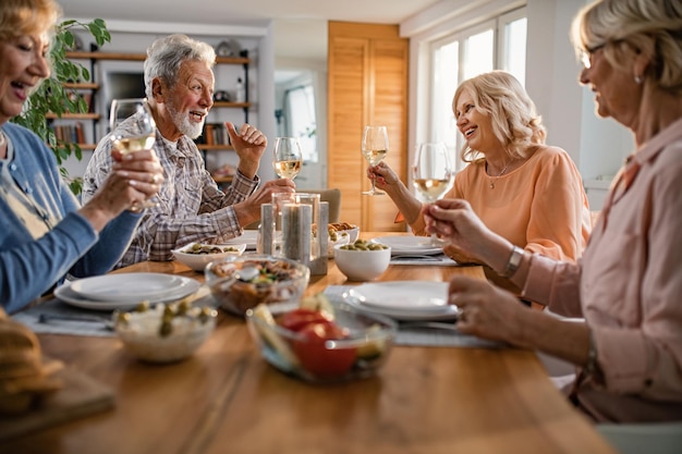 Groep gelukkige senioren roosteren met wijn tijdens het communiceren tijdens een lunch in de eetkamer