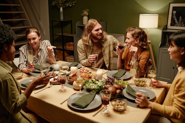 Groep gelukkige vrienden die aan tafel met elkaar praten en wijn drinken tijdens het etentje in de woonkamer