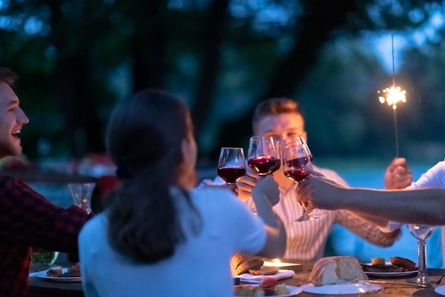 groep gelukkige vrienden die een glas rode wijn roosteren terwijl ze een picknick Frans etentje buiten hebben tijdens de zomervakantie in de buurt van de rivier in de prachtige natuur