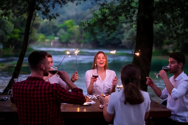 groep gelukkige vrienden die vakantie vieren met behulp van sproeiers en rode wijn drinken terwijl ze picknicken Frans etentje buiten in de buurt van de rivier op mooie zomeravond in de natuur