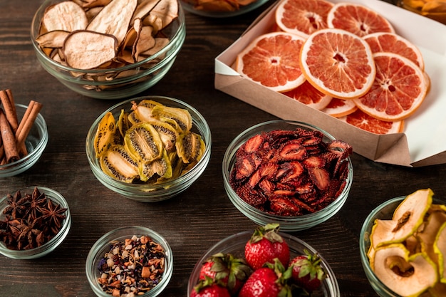 Groep glazen kommen met gedroogde kiwi, peren, aardbeien, sinaasappels en aromatische kruiden die op een donkere houten keukentafel staan