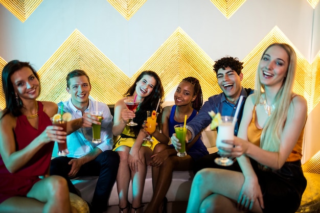 Groep glimlachende vrienden die op bank zitten en een glas cocktail tonen