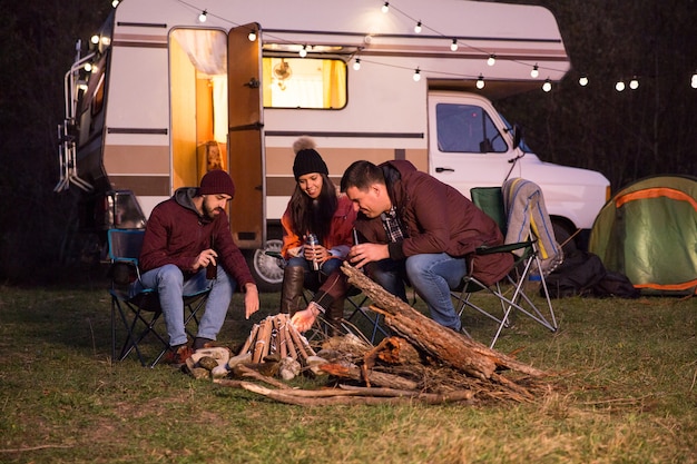 Groep goede vrienden die samen genieten van een biertje en kampvuur maken met hun retro camper op de achtergrond.