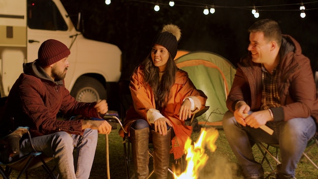 Groep goede vrienden die samen ontspannen rond het kampvuur in een koude herfstnacht. Retro camper. Kampeer tent.