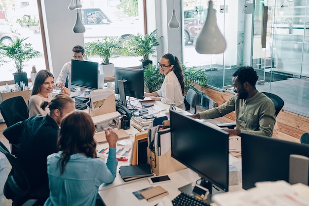 Groep jonge moderne mensen in slimme vrijetijdskleding die communiceren en moderne technologieën gebruiken terwijl ze op kantoor werken