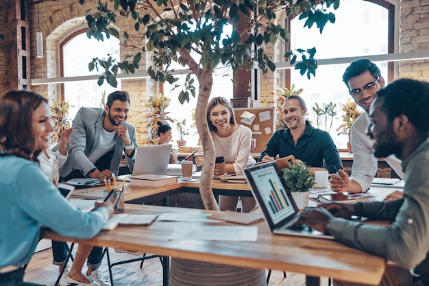 Groep jonge moderne mensen in slimme vrijetijdskleding die communiceren en moderne technologieën gebruiken terwijl ze op kantoor werken