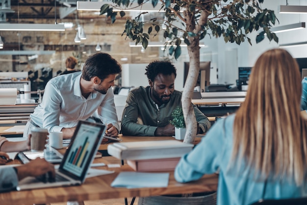 Groep jonge moderne mensen in slimme vrijetijdskleding die communiceren en moderne technologieën gebruiken terwijl ze op kantoor werken