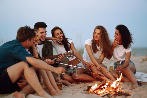 Groep jonge vrienden die op het strand zitten en worstjes bakken Een man speelt gitaar Kampeertijd