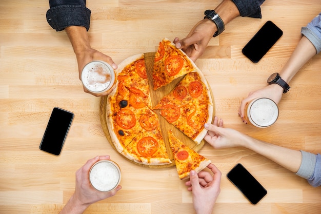 Groep jonge vrienden die pizza proeven en bier drinken op houten tafel