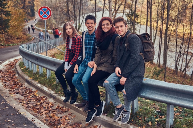 Groep jonge vrienden met rugzakken zittend op vangrail in de buurt van weg met een prachtig bos en rivier op de achtergrond. Reizen, wandelen, concept.