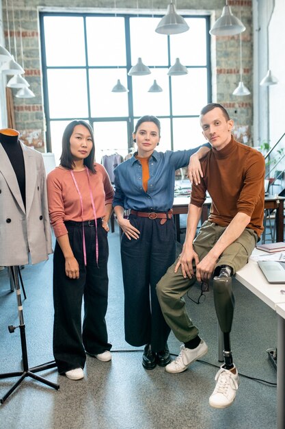 Groep jonge zelfverzekerde ontwerpers van kleding die voor de camera in de werkplaats staan