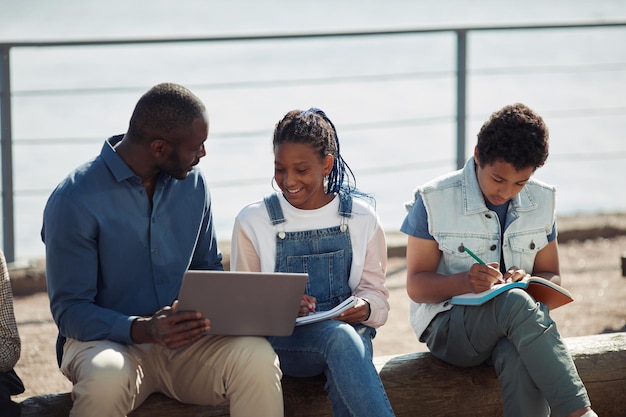 Groep lachende zwarte kinderen met leraar die laptop samen gebruikt tijdens buitenles in de zomerschool