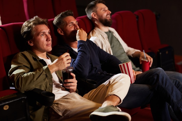 Groep mannelijke vrienden kijken naar film in bioscoop theater en popcorn eten zittend in rij op rode stoelen, kopieer ruimte