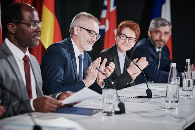 Foto groep mensen die de toespraak van hun collega applaudisseren terwijl ze aan tafel zitten tijdens de conferentie