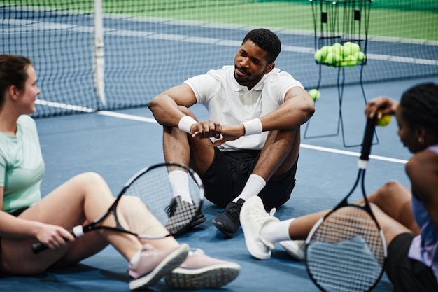 Groep mensen ontspannen op de indoor tennisbaan en kletsen terwijl ze op de vloer zitten