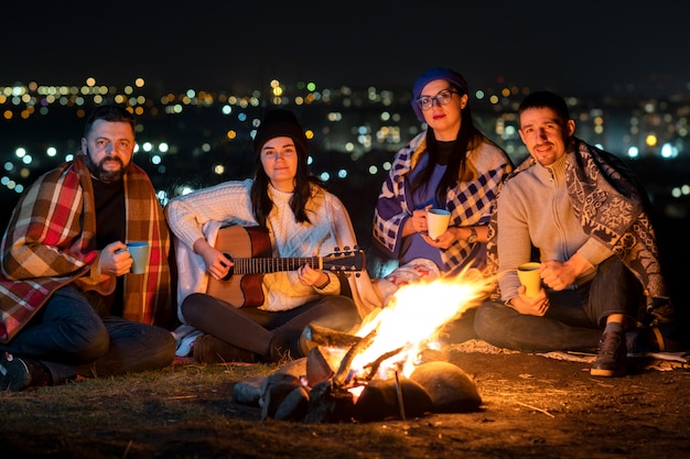 Groep mensen plezier zitten in de buurt van vreugdevuur buiten 's nachts gitaar spelen, liedjes zingen en gelukkig samen praten.