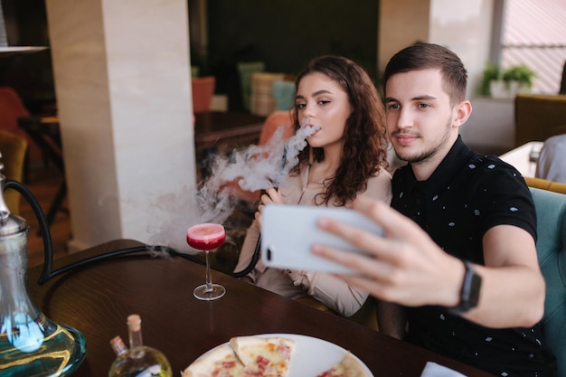 Groep mensen roken waterpijp op terras Man en vrouw ontmoeten elkaar in restaurant Man maakt verhalen terwijl zijn vriendin rookt Man maakt selfie Mooie mensen