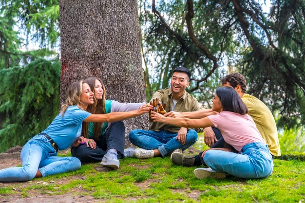 Groep multi-etnische vrienden zitten in het stadspark te praten naast een boom en roosteren met flesjes bier