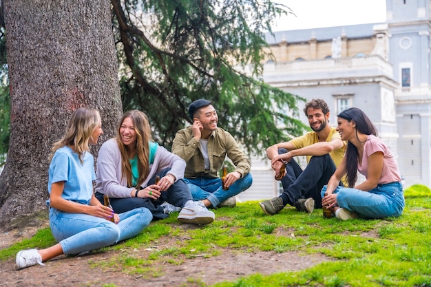 Groep multi-etnische vrienden zitten in het stadspark te praten naast een boom met flesjes bier