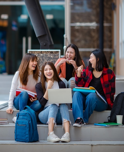 Groep opgetogen Aziatische vrouwelijke studenten die zich verzamelen op trappen op de universiteitscampus en samen huiswerk maken terwijl ze een laptop gebruiken. Concept voor goede vriend van het tienerleven.