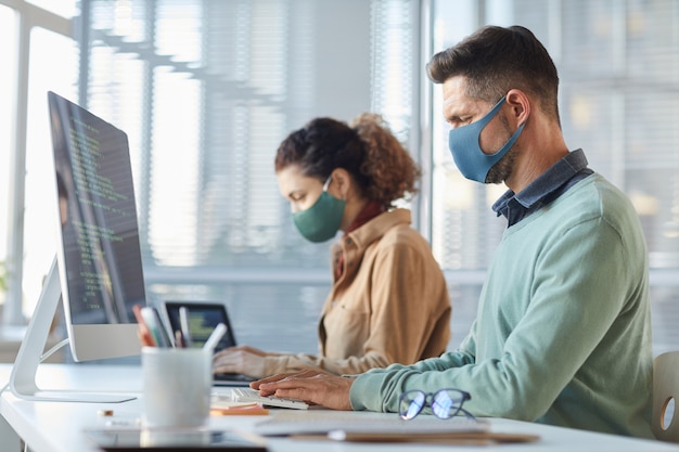 Groep programmeurs in maskers aan tafel zitten en werken met codes op computers tijdens pandemie