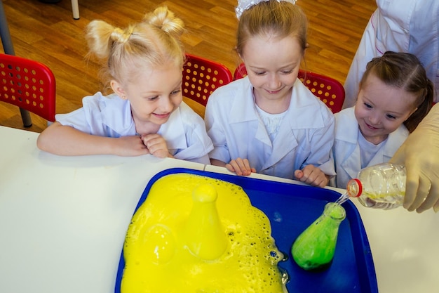 Groep schoolmeisjeskinderen met leraar in schoollaboratorium die experiment maken en de chemische reactie observeren met de kleurstof met azijn en soda-vulkaan die witte jurk uniform glas draagt