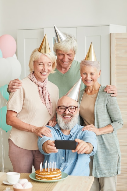 Groep senior vrienden in hoeden glimlachen naar de camera tijdens het maken van selfie op mobiele telefoon tijdens verjaardagsfeestje