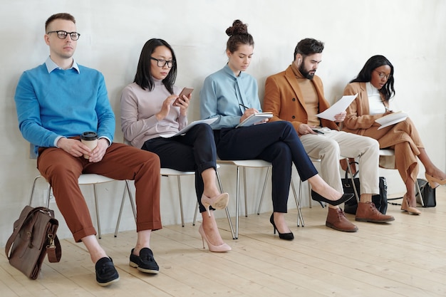 Groep serieuze jonge multi-etnische sollicitanten in slimme casual outfits die op stoelen in de rij zitten en zich voorbereiden op een interview in de gang