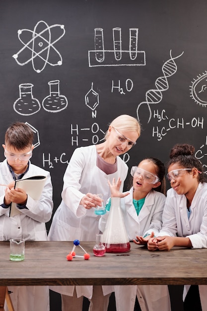Groep slimme leerlingen van de middelbare school in witte jassen en brillen die naar hun leraar kijken die een chemisch experiment laat zien op schoolbord