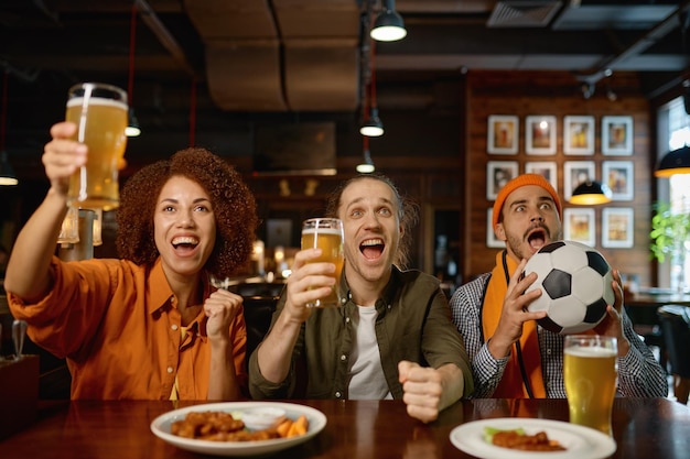 Groep sportfans die het doel vieren voor het favoriete voetbalteam. Jonge diverse multiraciale juichende mensen die ambachtelijk bier drinken terwijl ze naar een voetbalwedstrijd kijken in de sportbar