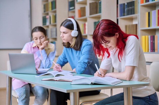 Groep tienerstudenten studeert aan hun bureau in de bibliotheekklas Tienerstudenten schrijven in notitieboeken met behulp van boeken laptop Onderwijs kennis middelbare school college concept