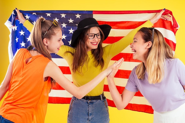 Groep van drie Aziatische en Europese lachende meisjes met paspoorten en kaartjes fotocamera verpakt in Amerikaanse vlag lentefeest zomer stijl gele achtergrond studiovisa en onafhankelijkheidsdag usa