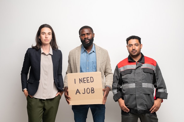 Groep van drie jonge interculturele werkloze mensen die zich door muur voor camera bevinden terwijl één van hen nota over behoefte aan baan vasthoudt