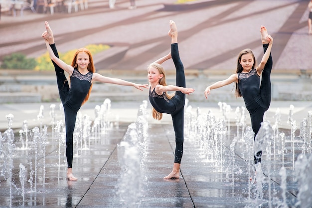 Groep van drie kleine ballerina's in zwarte strakke pakken dansen op een warme dag tegen de achtergrond van stadsfonteinen.