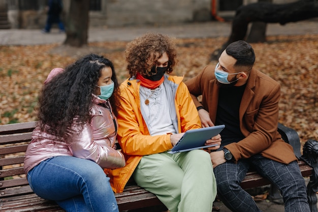 Groep van drie multiculturele mensen in medische maskers die op bank zitten en digitale tablet gebruiken. Jonge mannelijke en vrouwelijke vrienden ontspannen in herfst park.