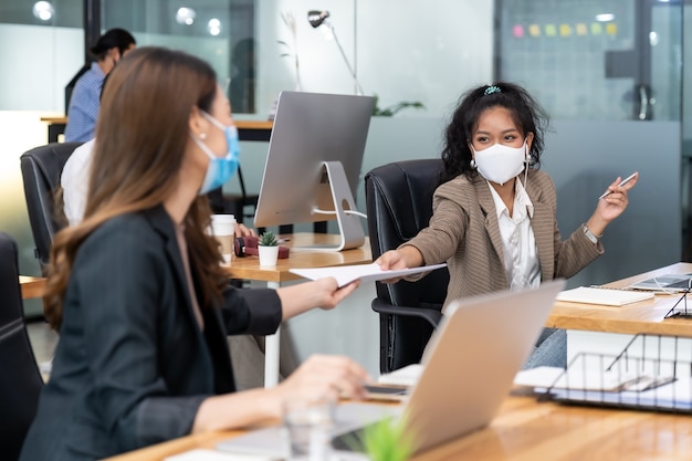 Groep van interraciale bedrijfsmedewerkers draagt een beschermend gezichtsmasker in een nieuw normaal kantoor en oefent op sociale afstand met handdesinfecterend alcoholgel op tafel om verspreiding van coronavirus COVID-19 te voorkomen