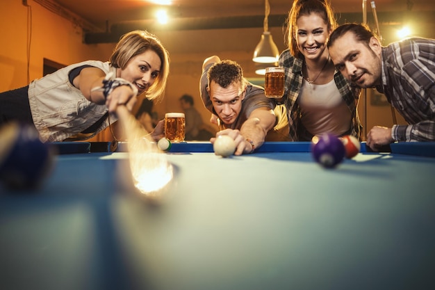 Groep van vier lachende vrolijke vrienden spelen biljart in de bar na het werk. Ze zijn betrokken bij recreatieve activiteiten.