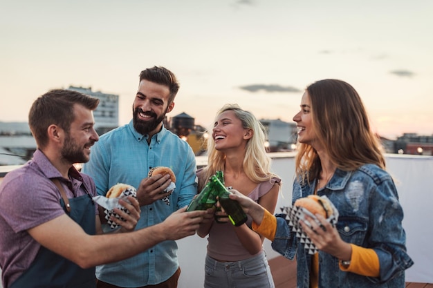 Groep vrienden die hamburgers eten en juichen met bier op het dak van een gebouw