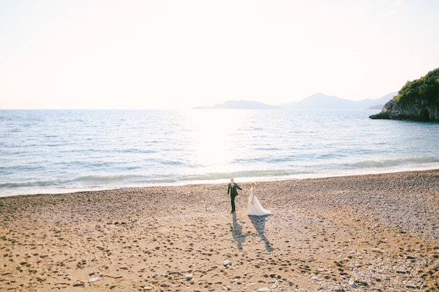 사진 하얀 드레스를 입은 신랑과 신부가 손을 잡고 꽃다발을 들고 해변을 걷고 있다